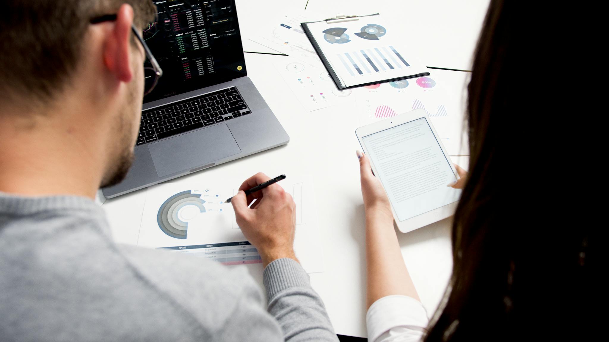 About Two business professionals analyzing financial reports using a laptop and tablet in a modern office setting.